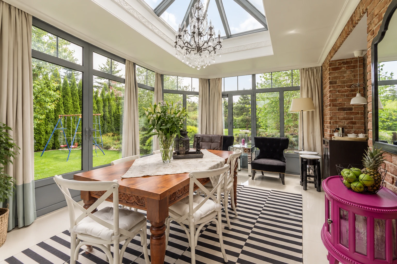 A bright four-season sunroom features floor-to-ceiling windows, a glass roof, dining furniture, and a brick accent wall.