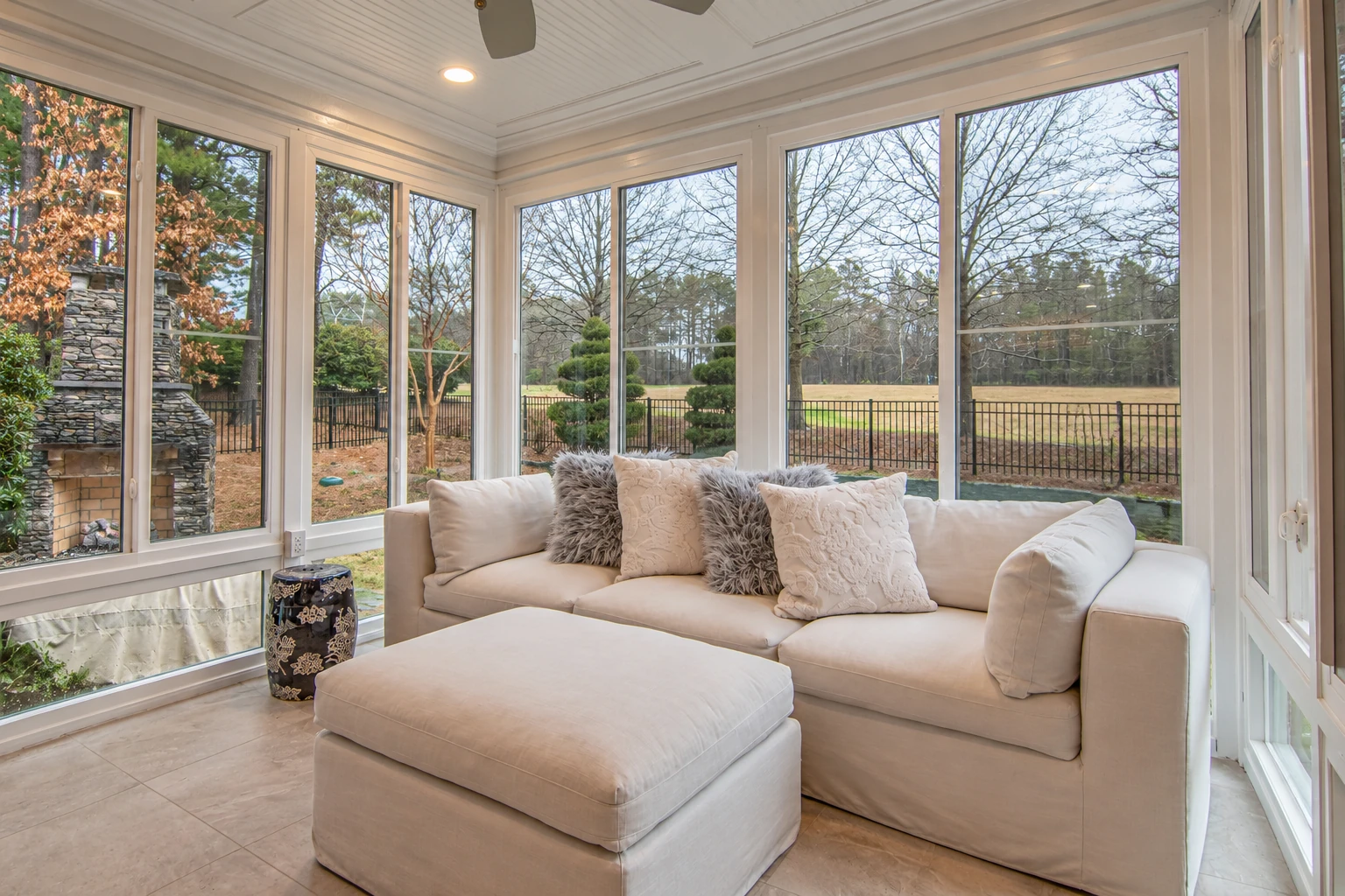 A sunroom with white-framed windows surrounds a cream sofa and ottoman, overlooking a fenced yard and trees.