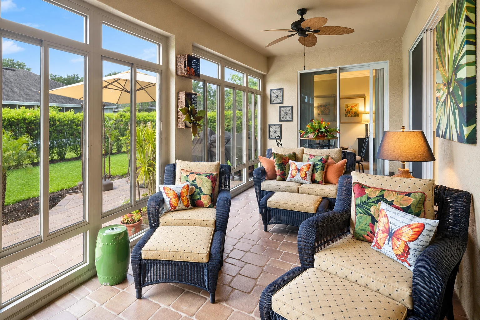 An enclosed patio room has sliding glass walls, tiled flooring, and wicker seating with colorful butterfly cushions.