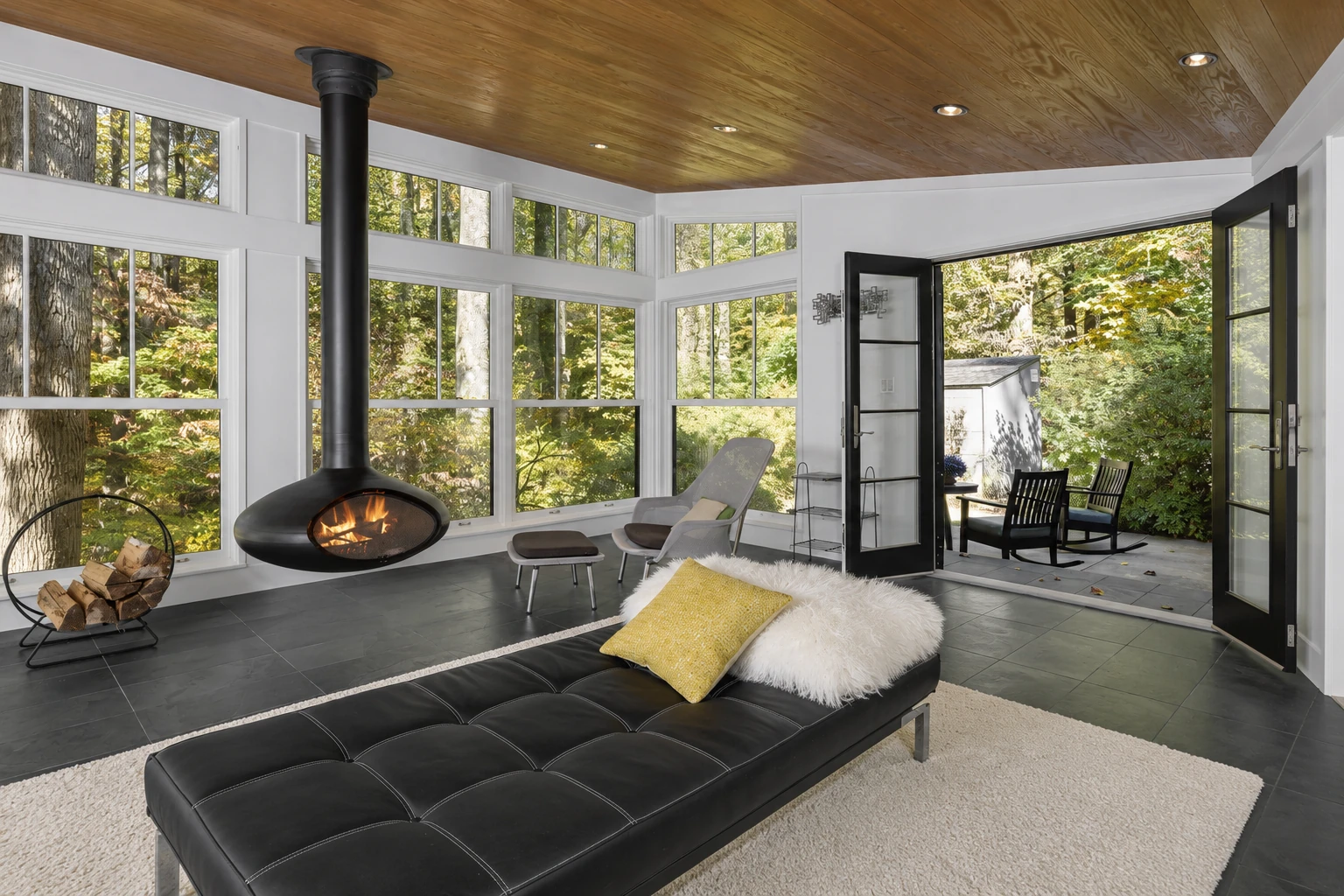 Four-season sunroom with a wood ceiling, large windows, a hanging fireplace, and open doors to a wooded patio.