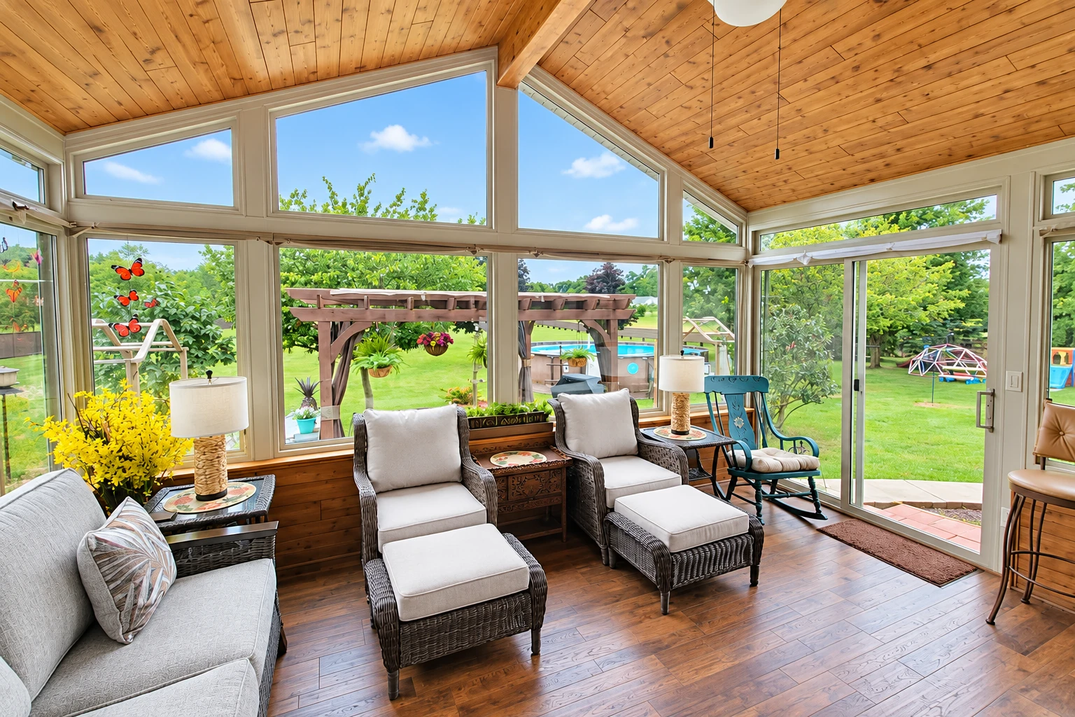 A bright four-season sunroom has a wood-paneled vaulted ceiling, wicker seating, and broad windows overlooking a green yard.