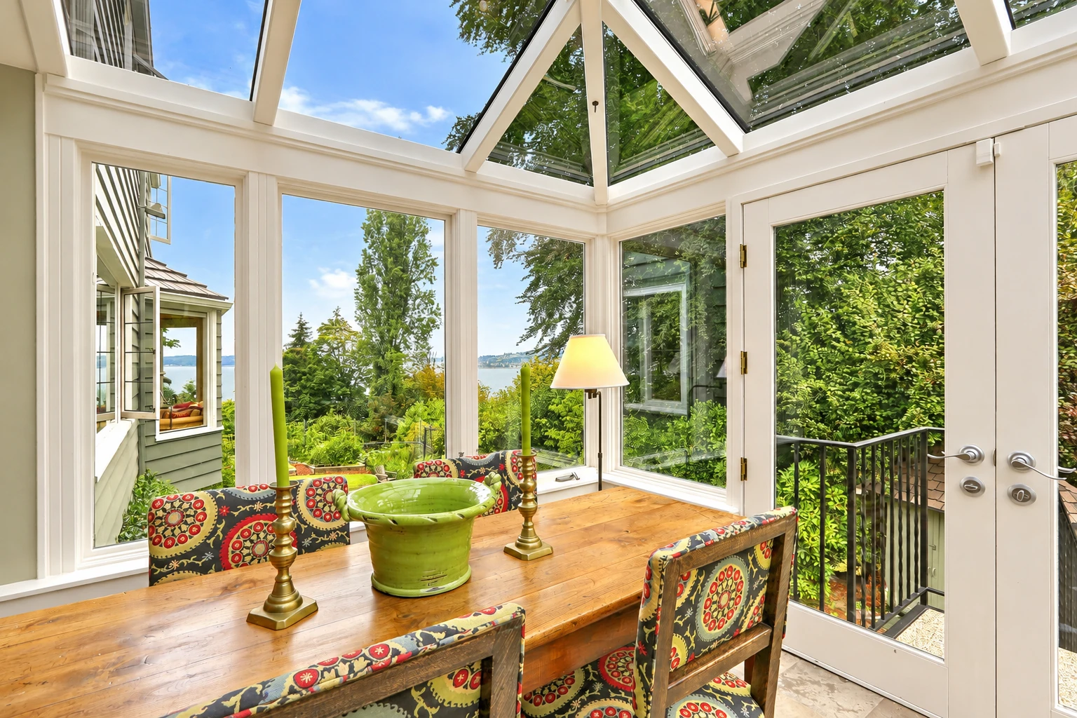 A glass-roofed solarium with white frames, a long wooden dining table, patterned chairs, and leafy views toward the water.