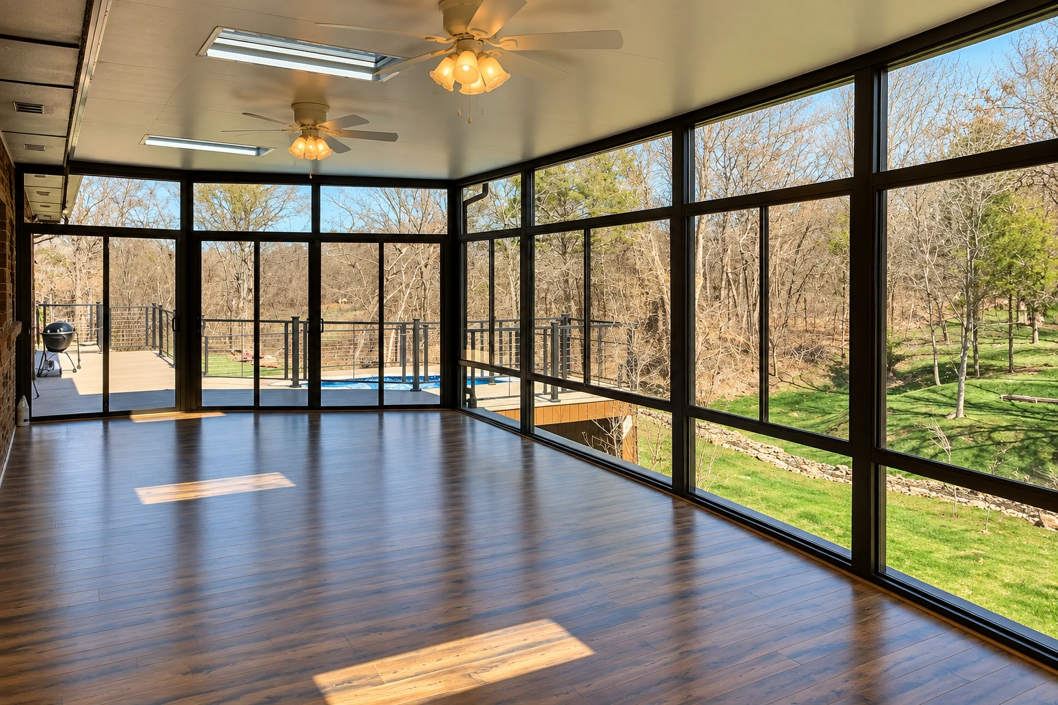 A spacious sunroom with dark-framed windows, ceiling fans, and a polished wood floor overlooking a deck and garden.