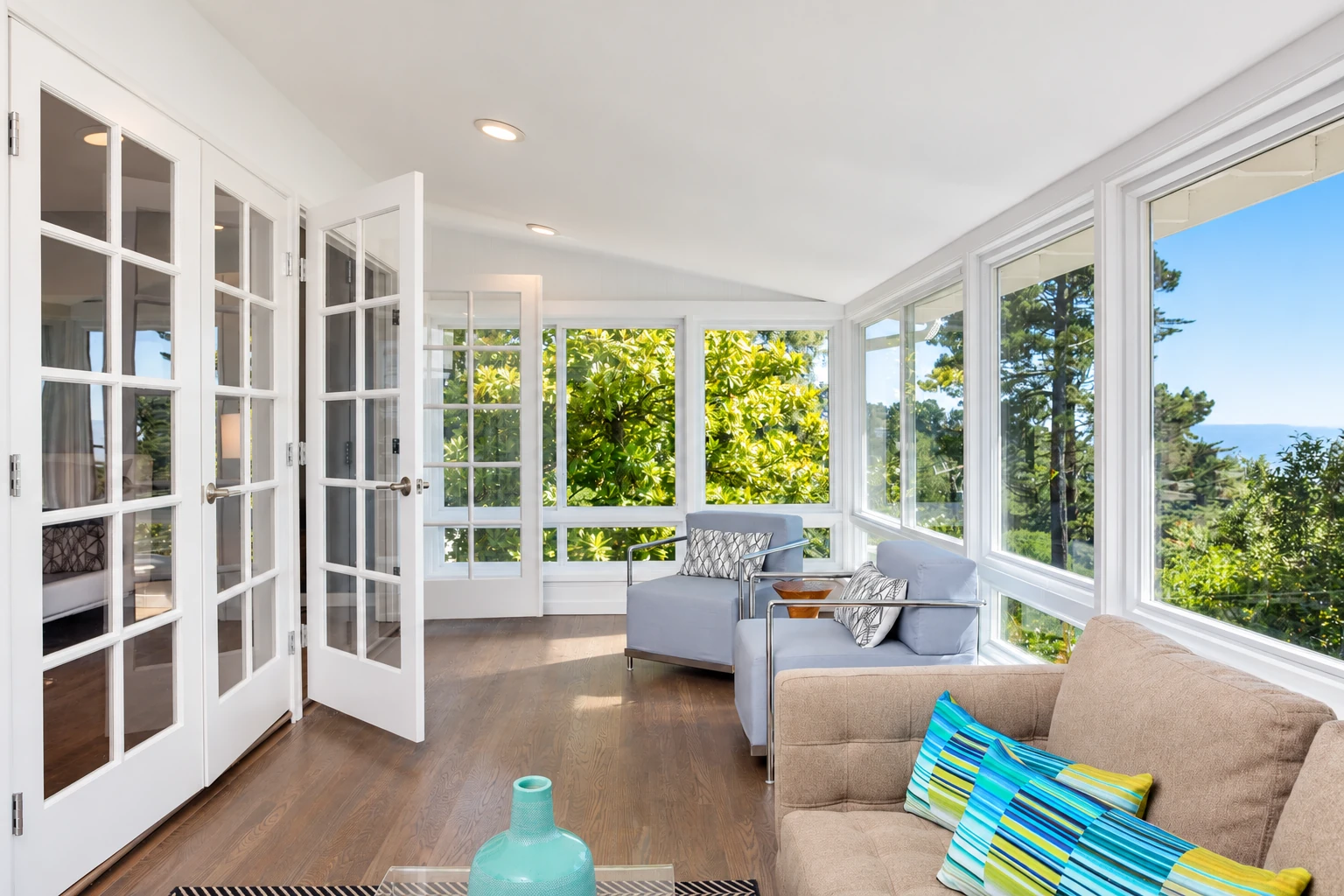 A bright remodeled sunroom with white-framed windows, open French doors, and pale seating surrounded by garden views.