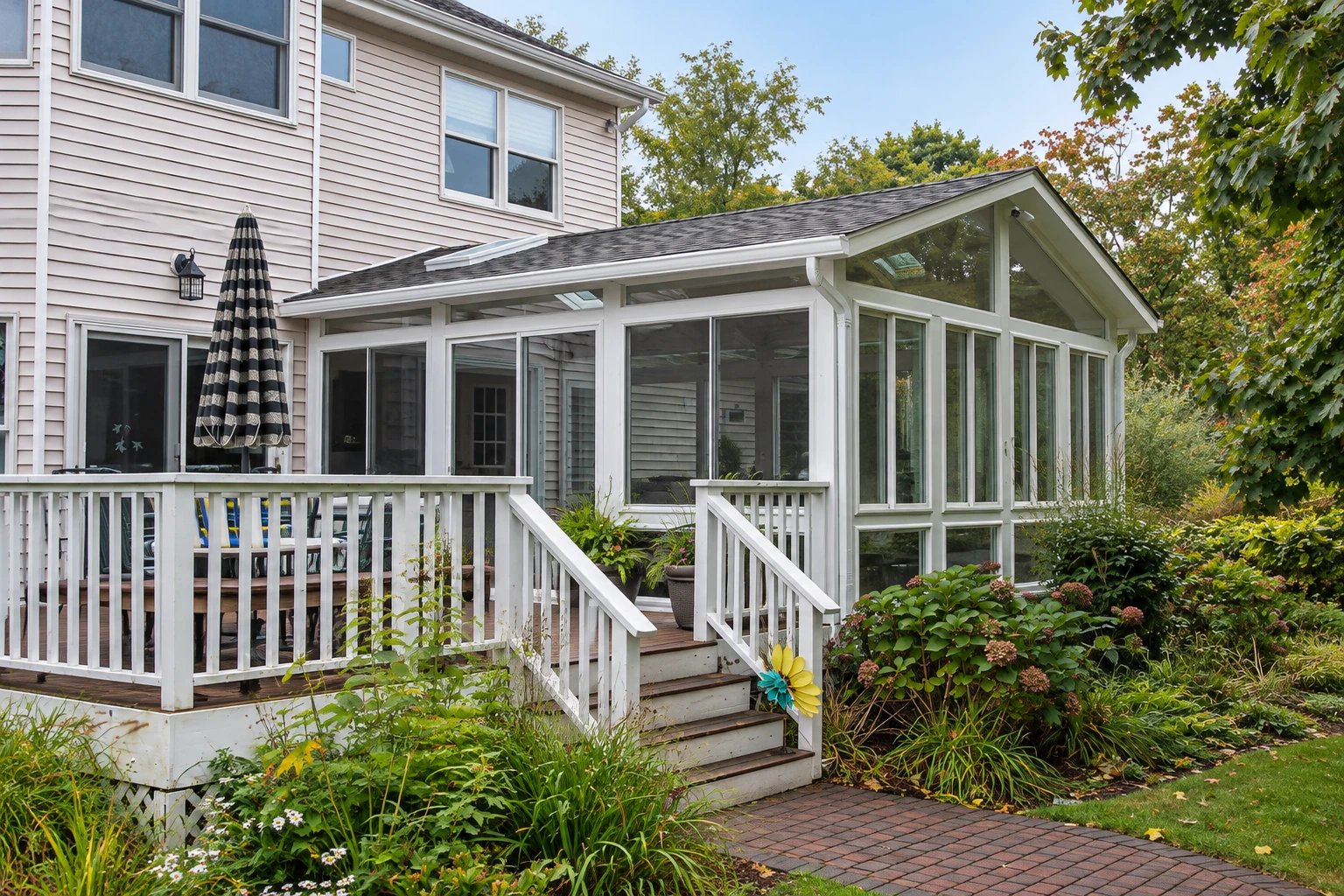 White-framed three-season sunroom extending from a house, with a gabled roof, deck steps, and garden beds.