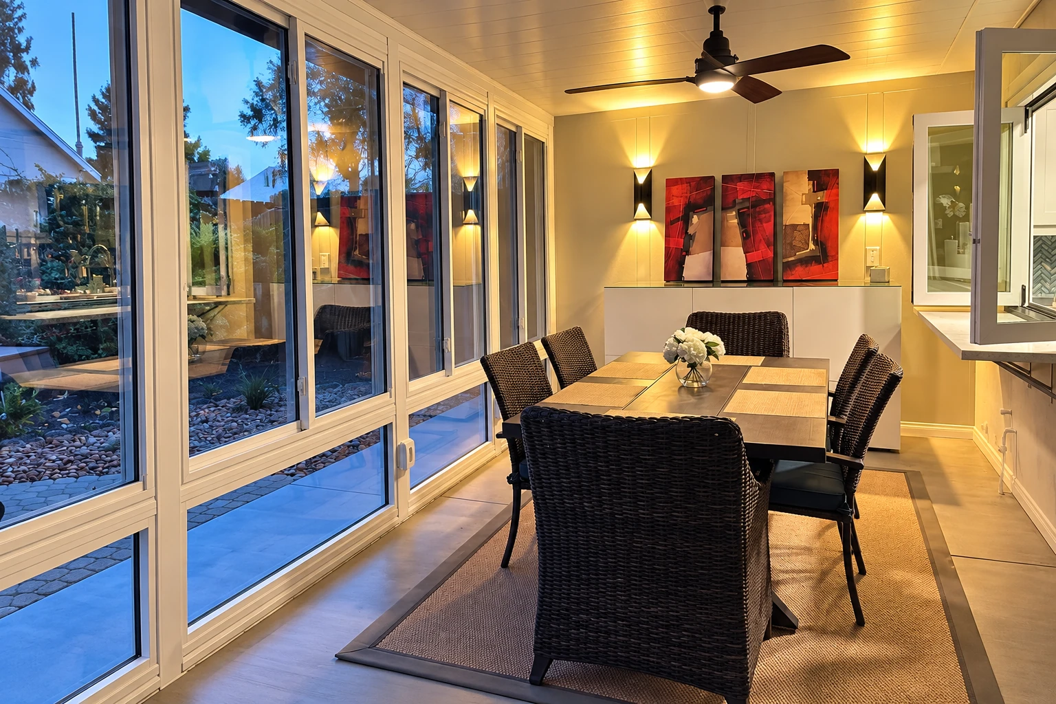 A three-season sunroom with tall windows, a dining table, dark wicker chairs, and warm evening lighting.