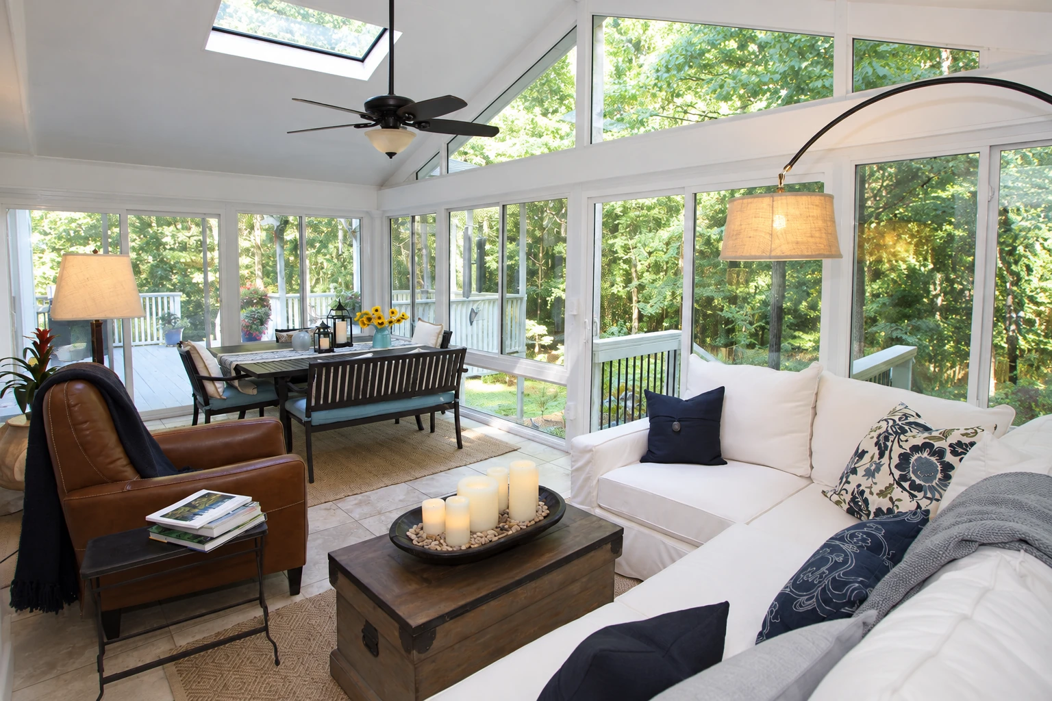 A bright vinyl sunroom combines a white sectional seating area with a dining table, surrounded by windows and leafy views.